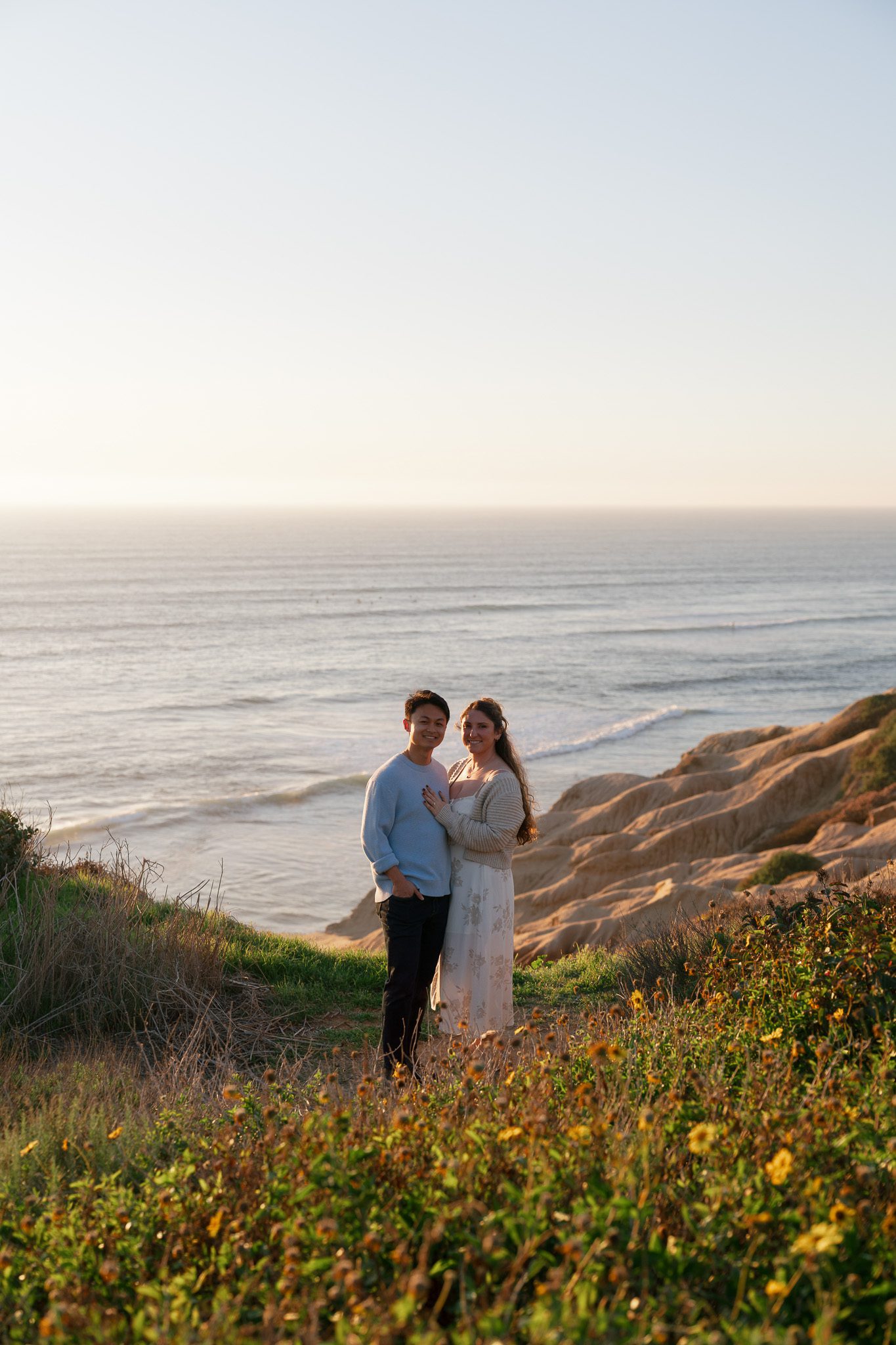 a couple posing in a field of wildflowers with the ocean in the background at sunset cliffs natural park, one of the best engagement photo locations in southern california.
