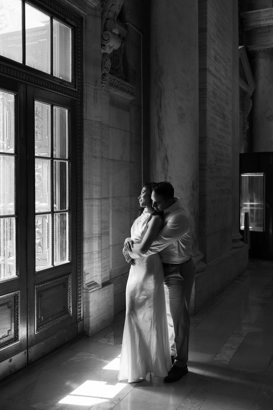 couple hugging in front of a window with lots of natural light pouring in during their NYC engagement photos. 